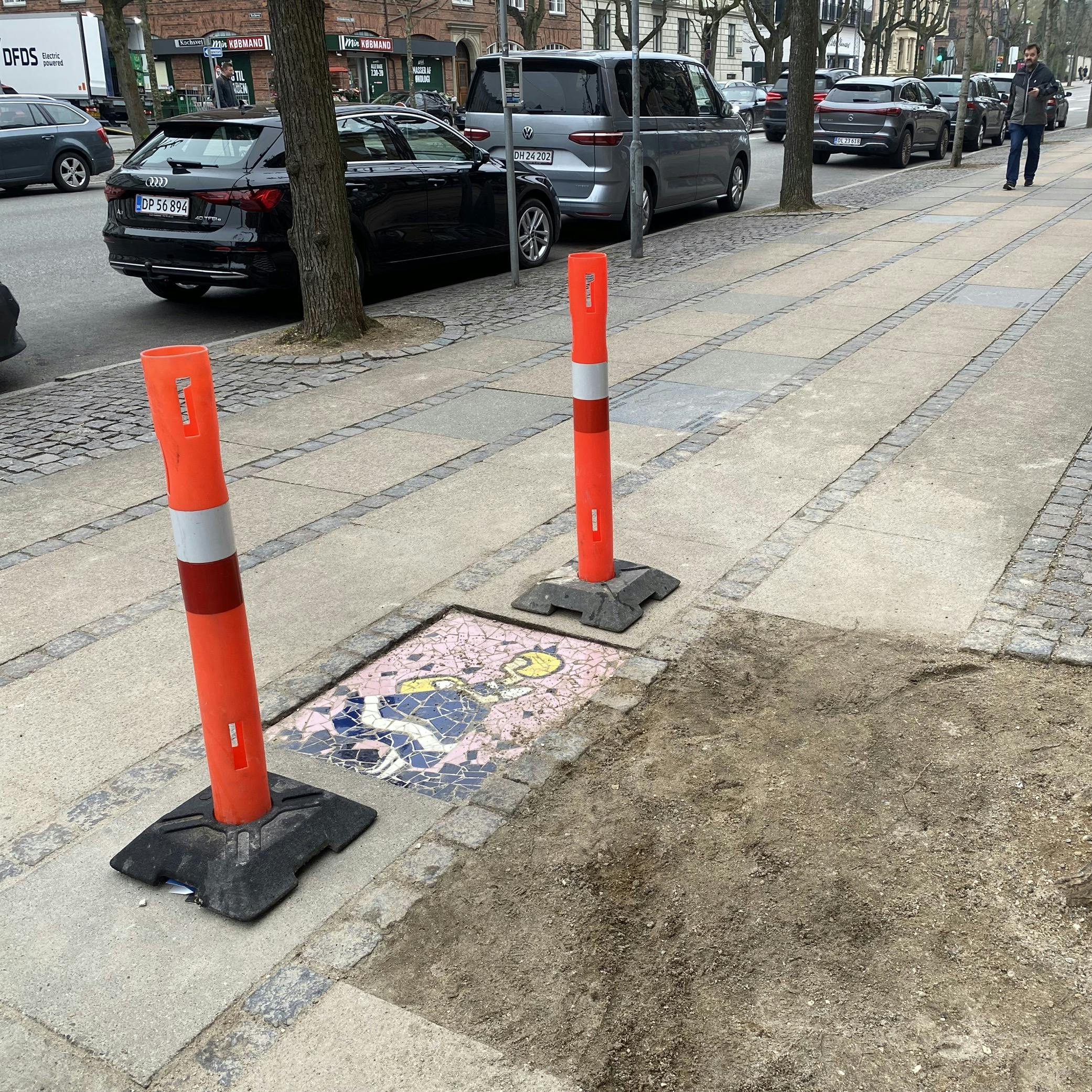 Mandag morgen har by-, kultur-, og miljøforvaltningen sat afspærringscylindre op ved mosaikflisen - nu skal lokalpolitikerne afgøre, om den skal blive liggende. 