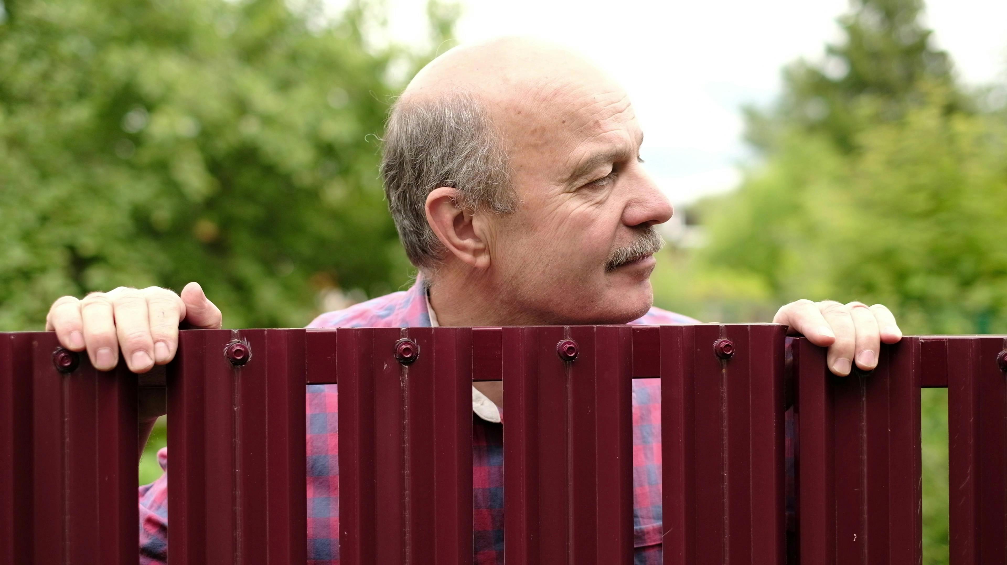 Mature caucasian man carefully watching over the fence. Concept of curious neighbors and private life
