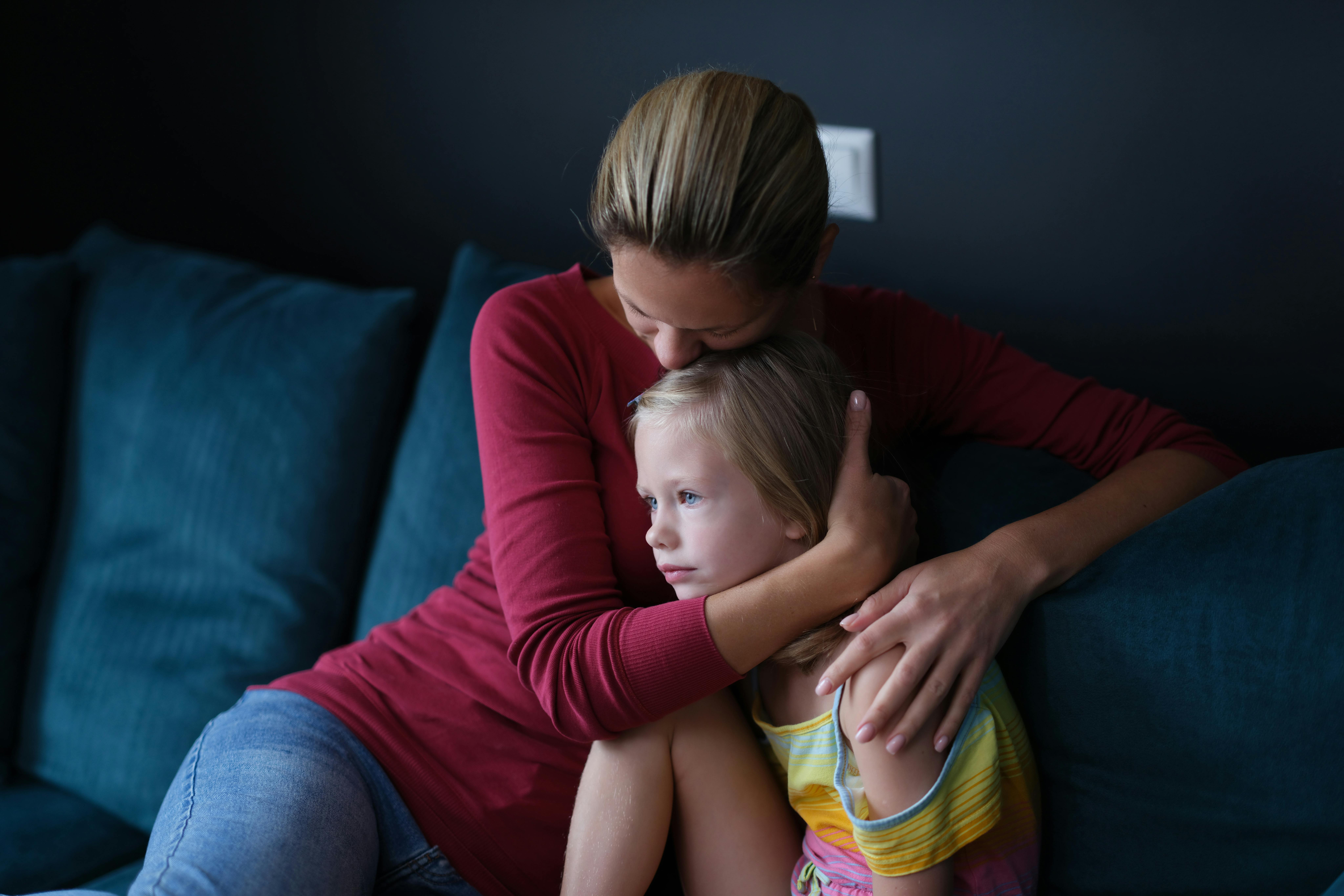 Woman and sad little daughter are sitting on couch and hugging. Maternal support for children concept