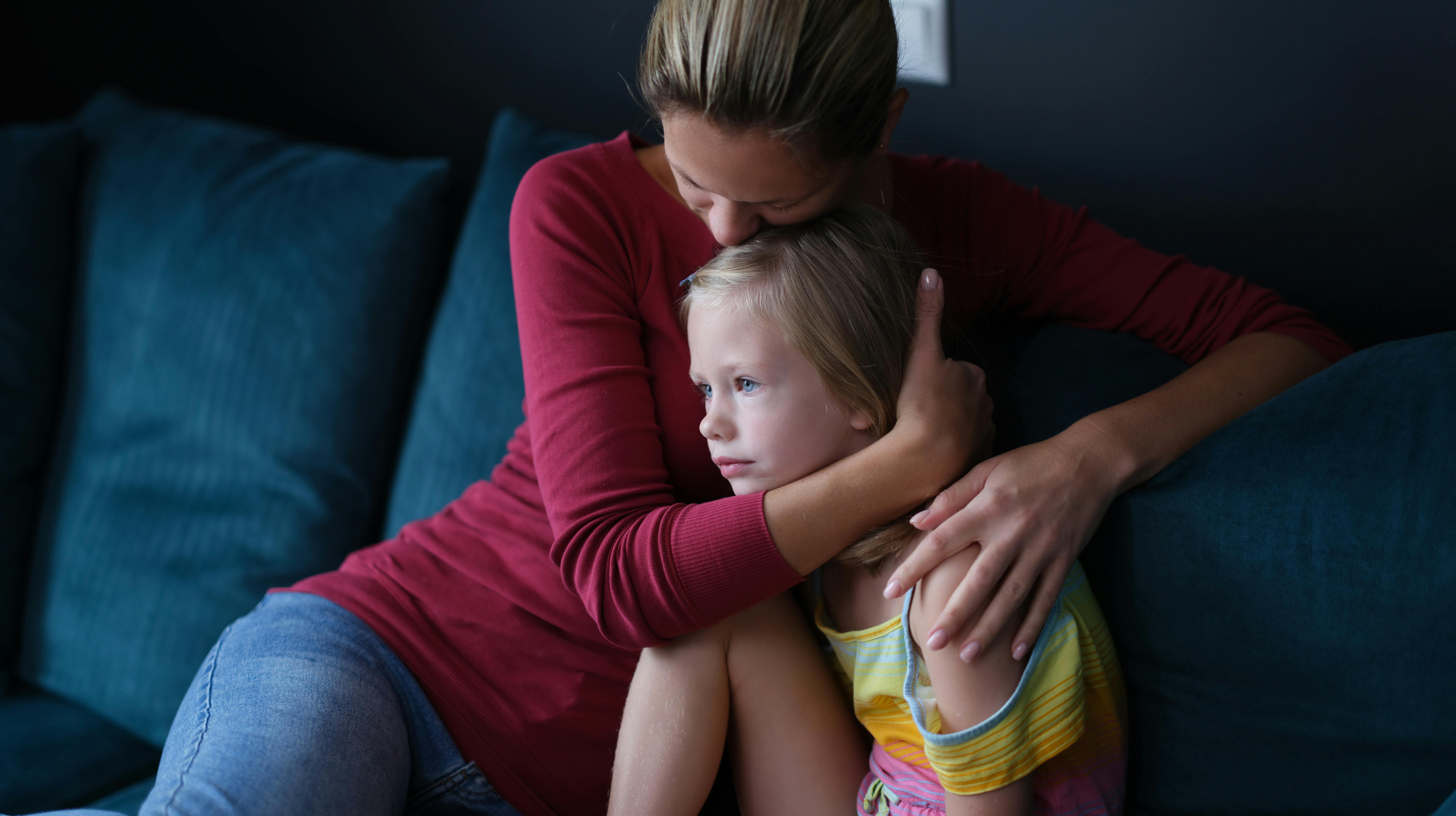 Woman and sad little daughter are sitting on couch and hugging. Maternal support for children concept