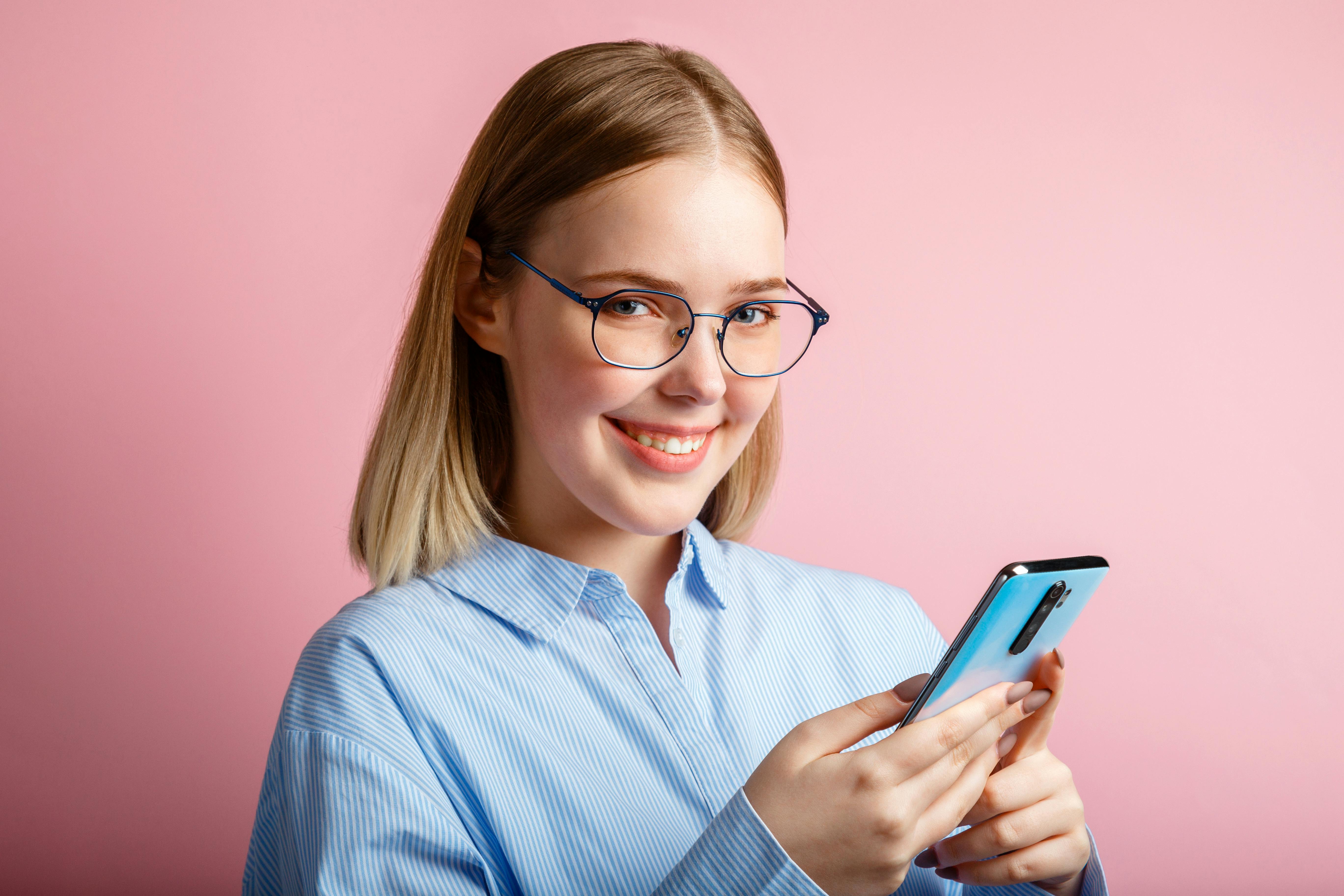 Emotional portrait of happy positive smile manager office worker in glasses use smartphone. Woman in a blue shirtwrites a message on phone isolated over color pink background