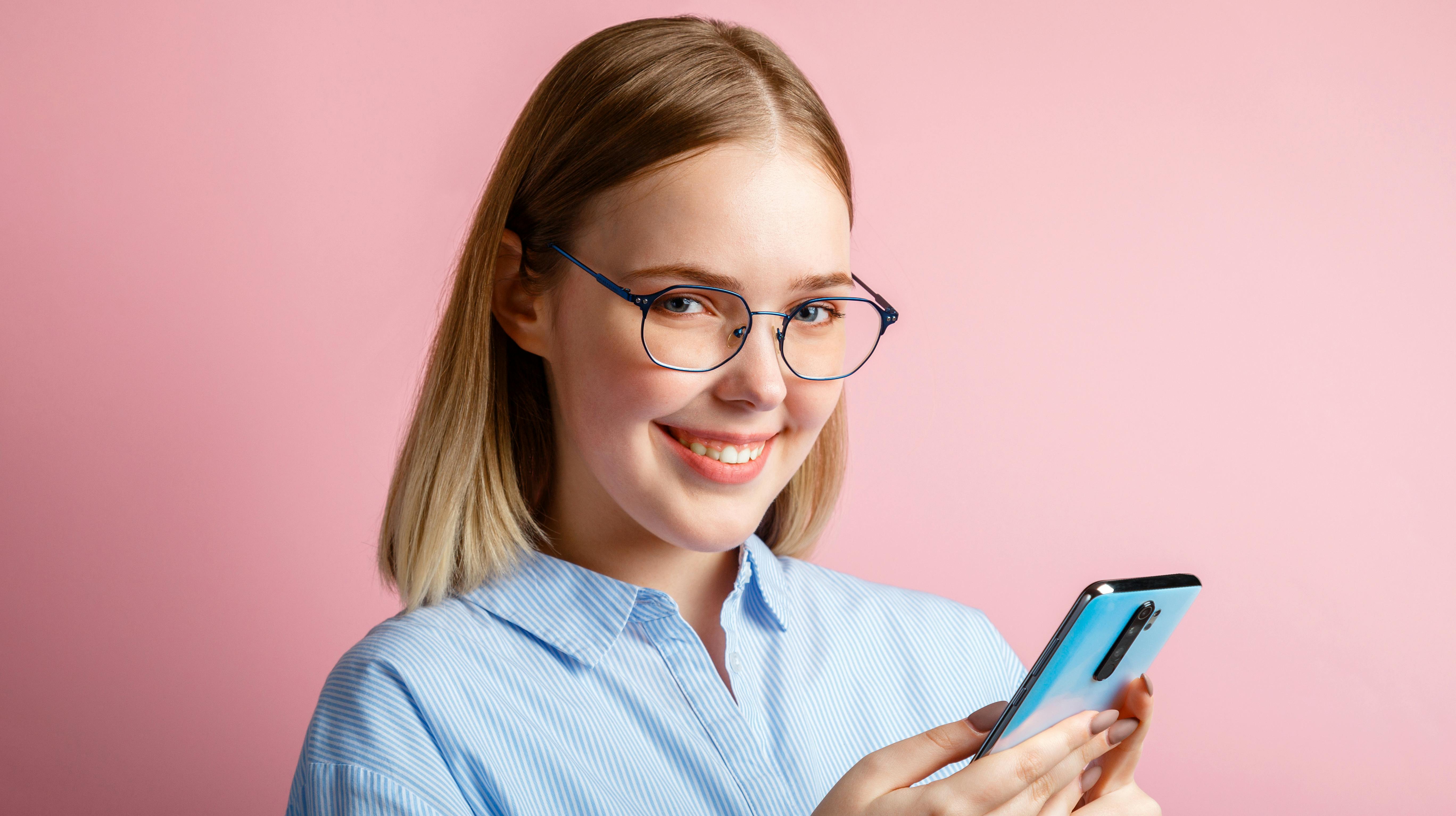 Emotional portrait of happy positive smile manager office worker in glasses use smartphone. Woman in a blue shirtwrites a message on phone isolated over color pink background