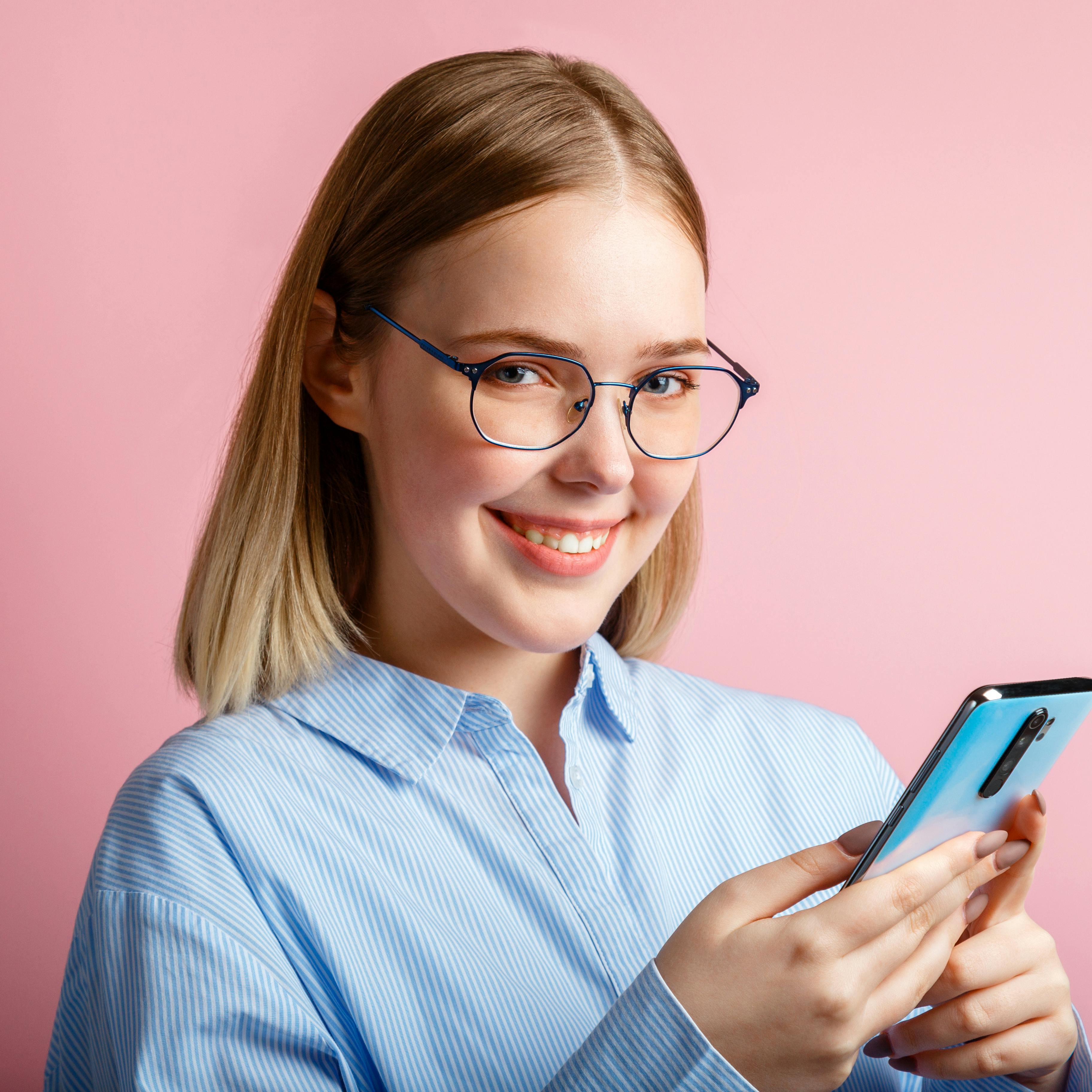 Emotional portrait of happy positive smile manager office worker in glasses use smartphone. Woman in a blue shirtwrites a message on phone isolated over color pink background