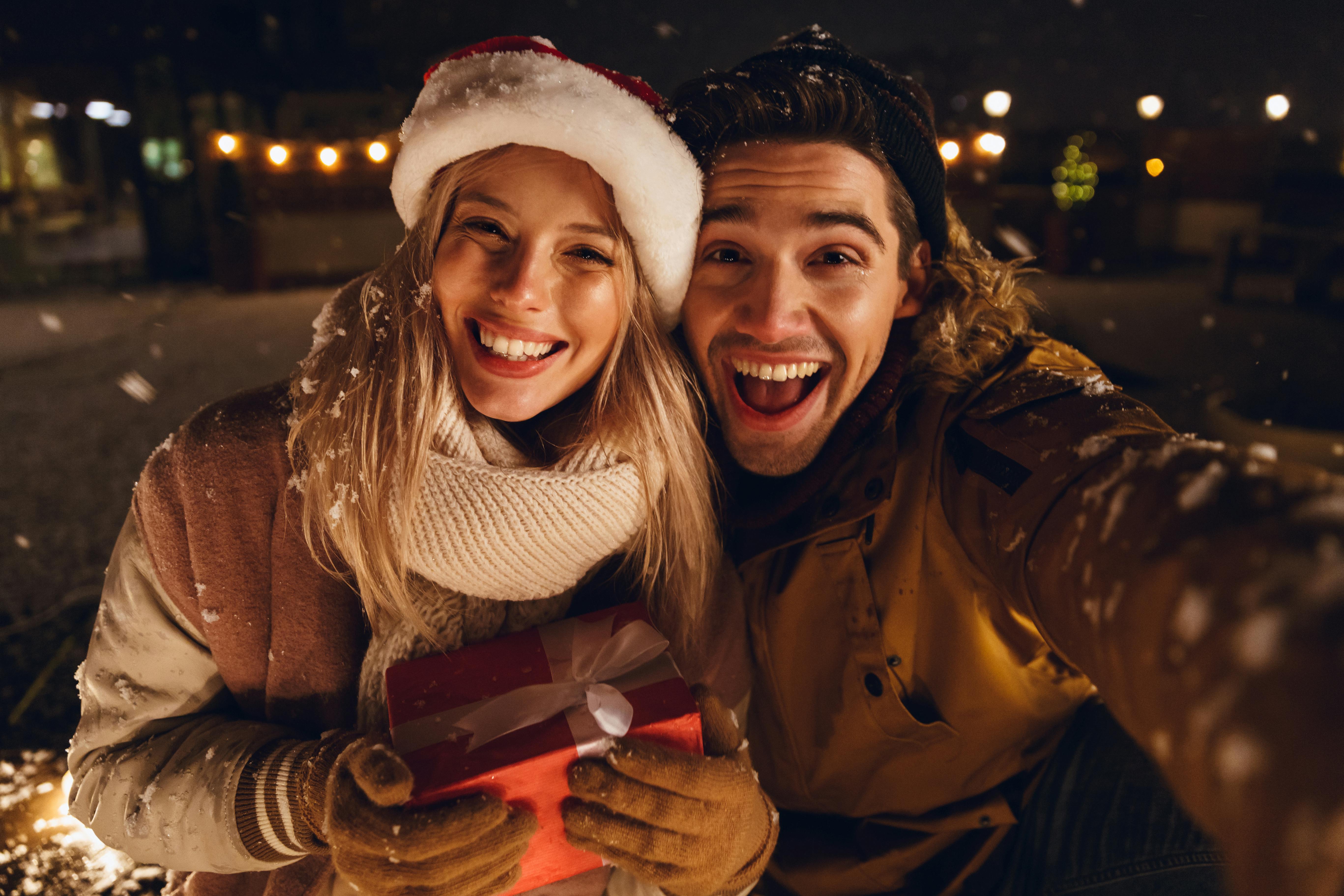 Photo of a happy young loving couple sitting outdoors in evening in christmas hat holding gift box take a selfie by camera.