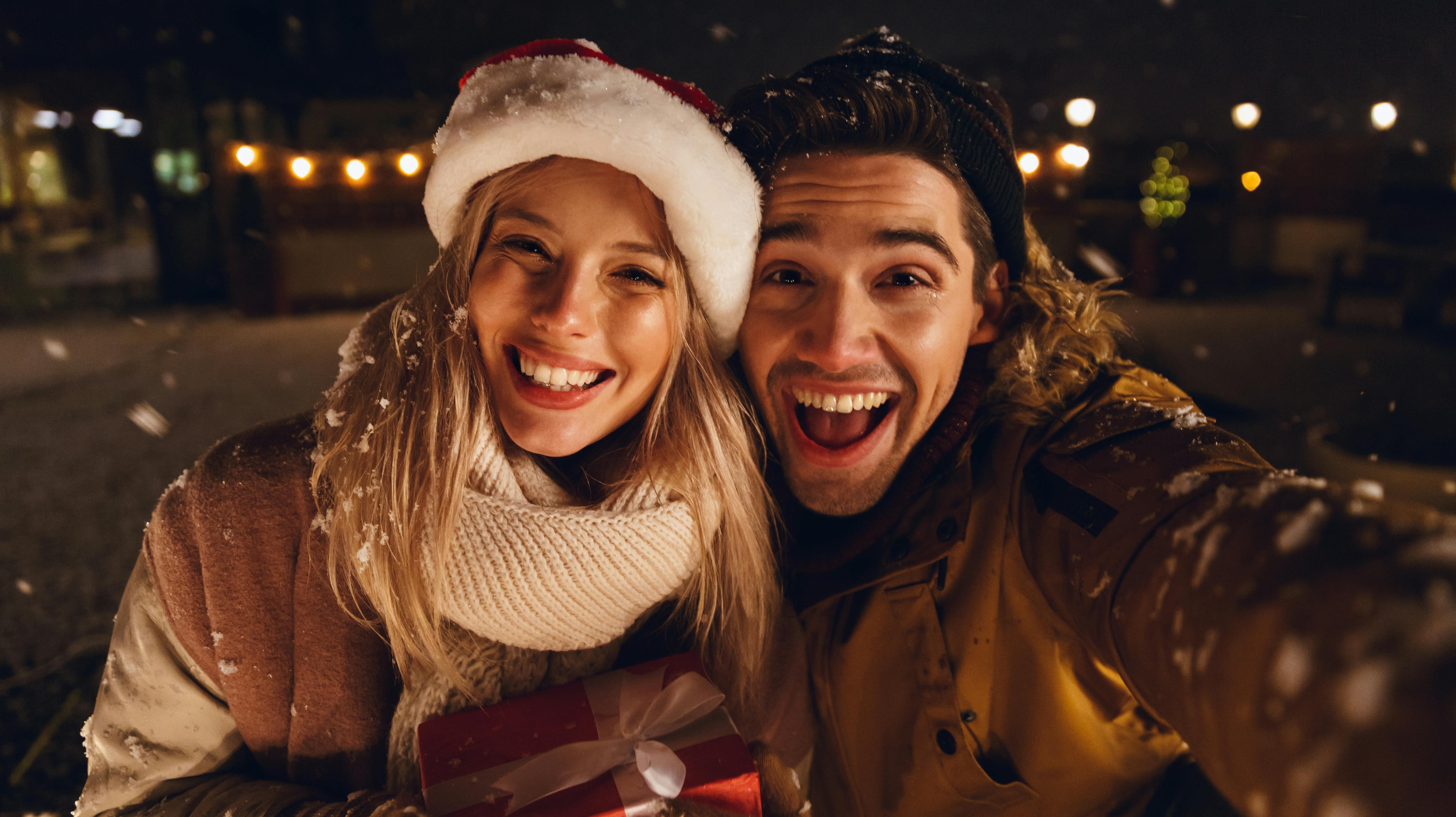 Photo of a happy young loving couple sitting outdoors in evening in christmas hat holding gift box take a selfie by camera.