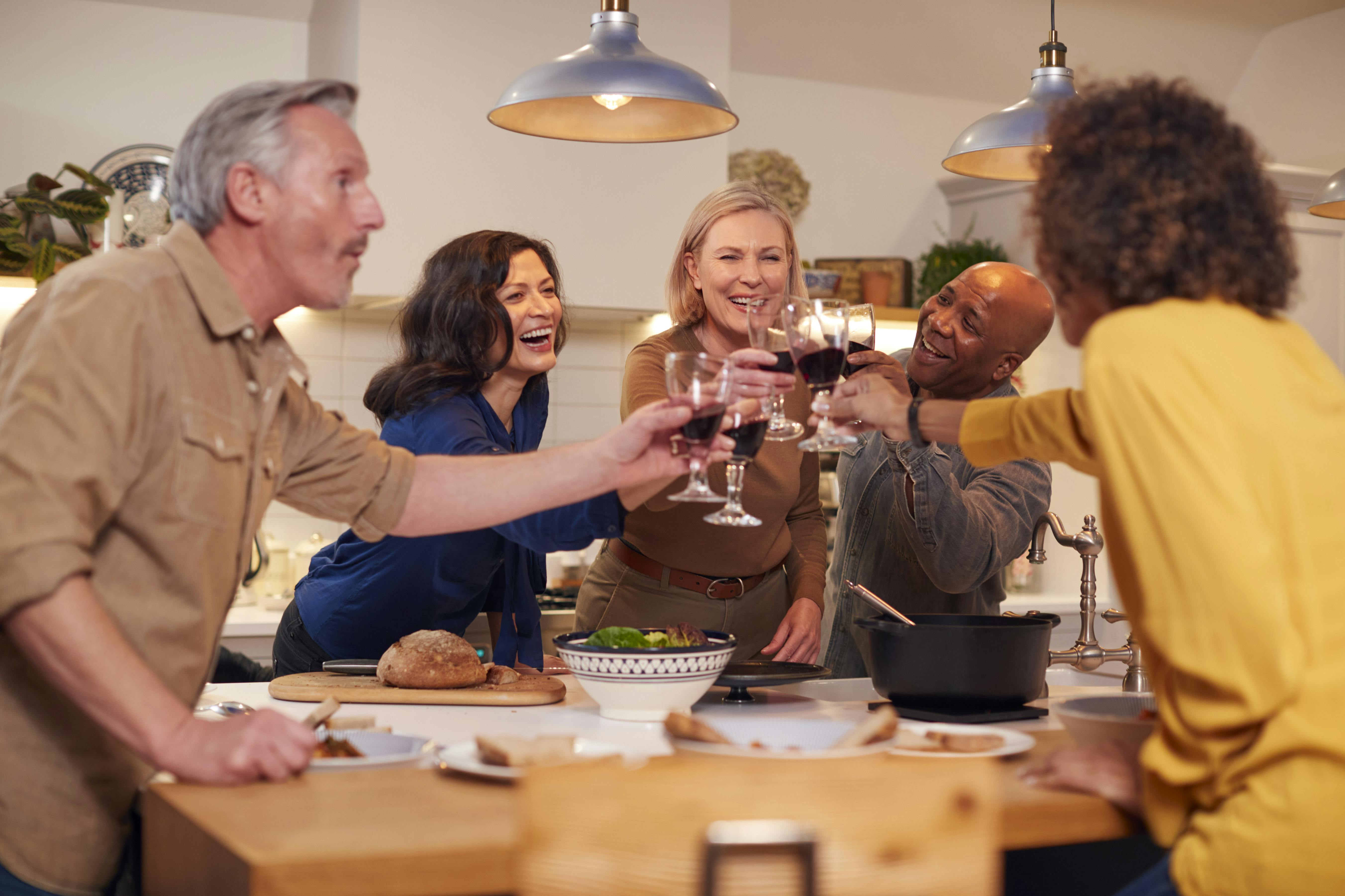 Group Of Mature Friends Making A Toast As They Meet At Home For Meal And Serve Food In Kitchen