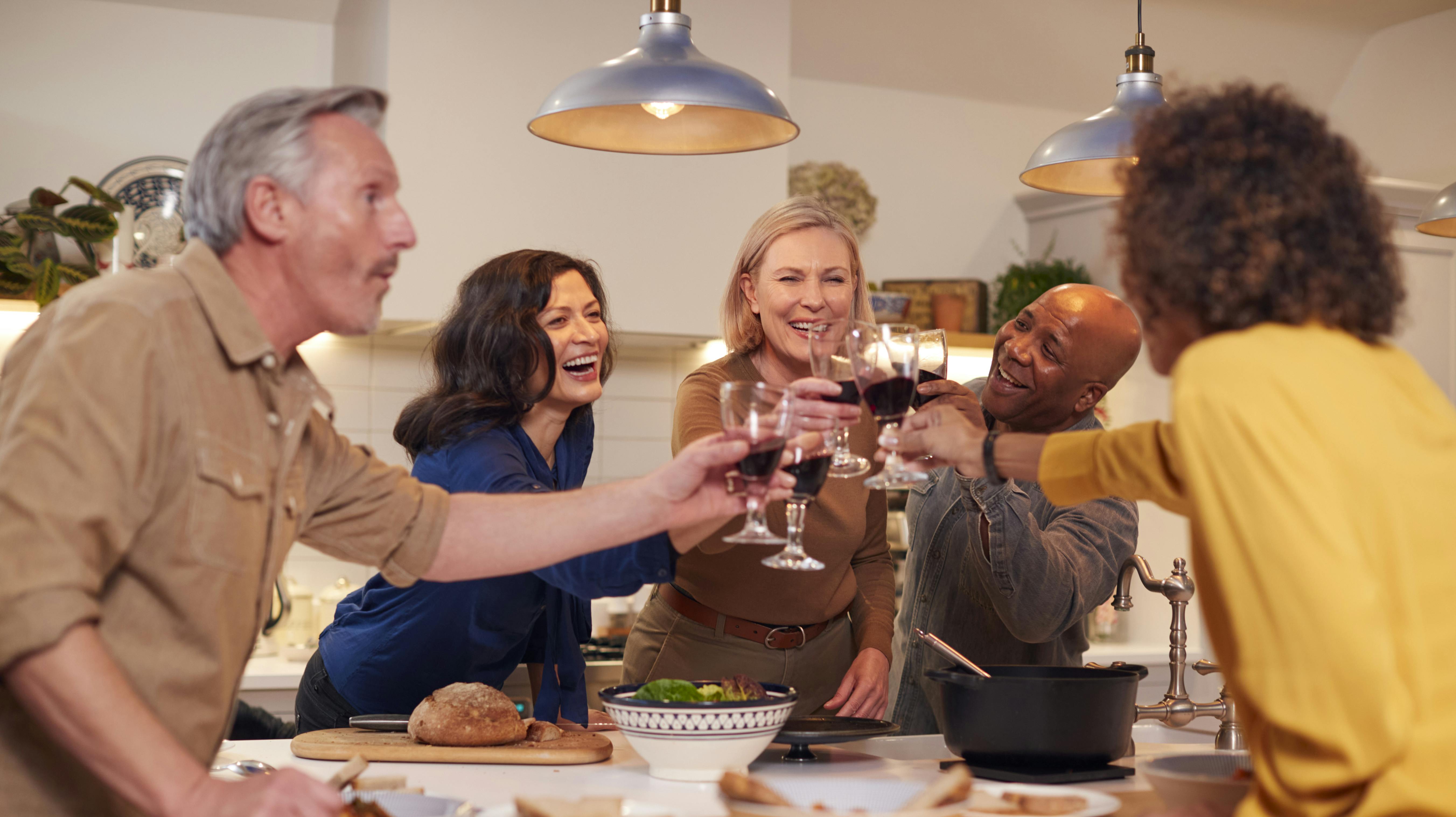 Group Of Mature Friends Making A Toast As They Meet At Home For Meal And Serve Food In Kitchen