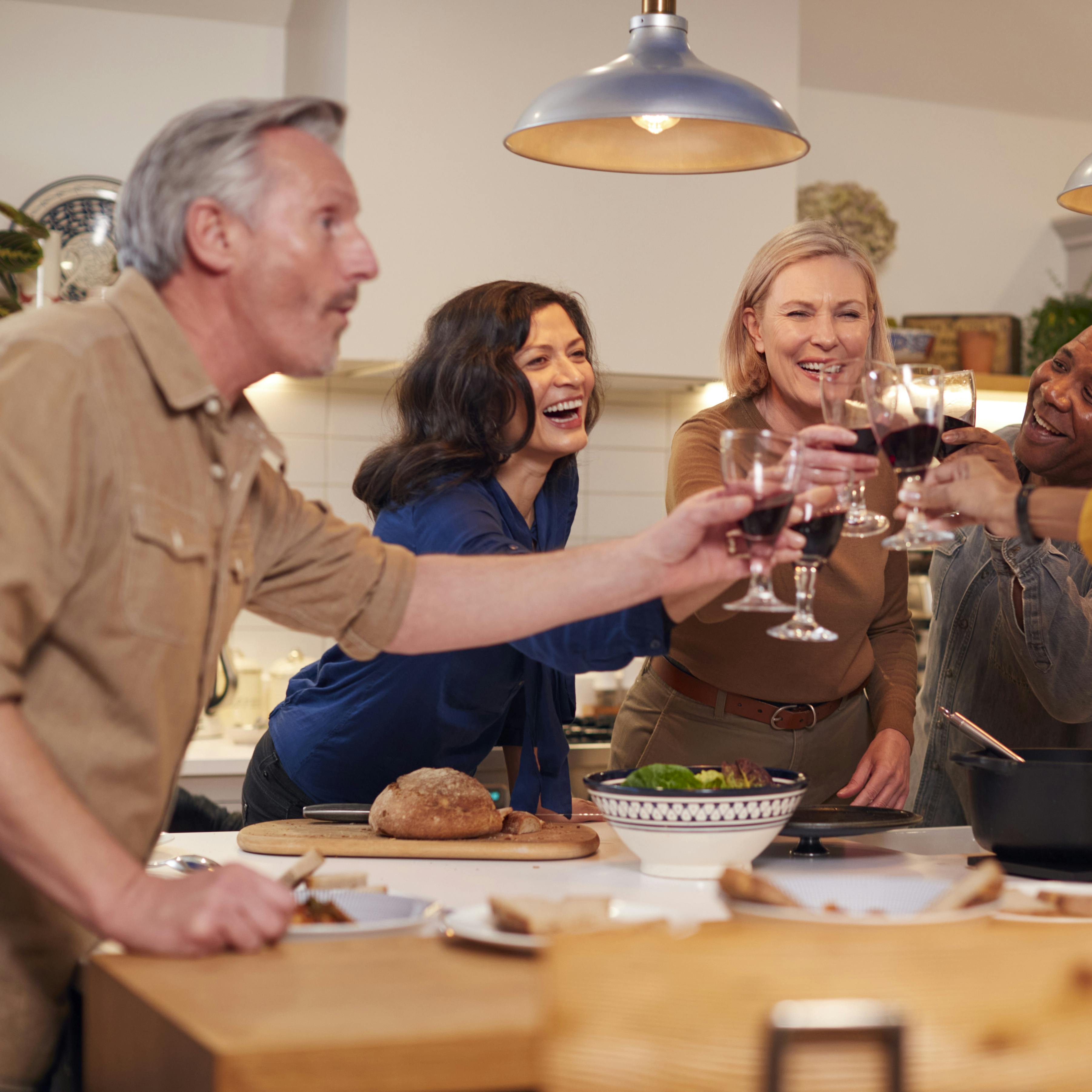 Group Of Mature Friends Making A Toast As They Meet At Home For Meal And Serve Food In Kitchen