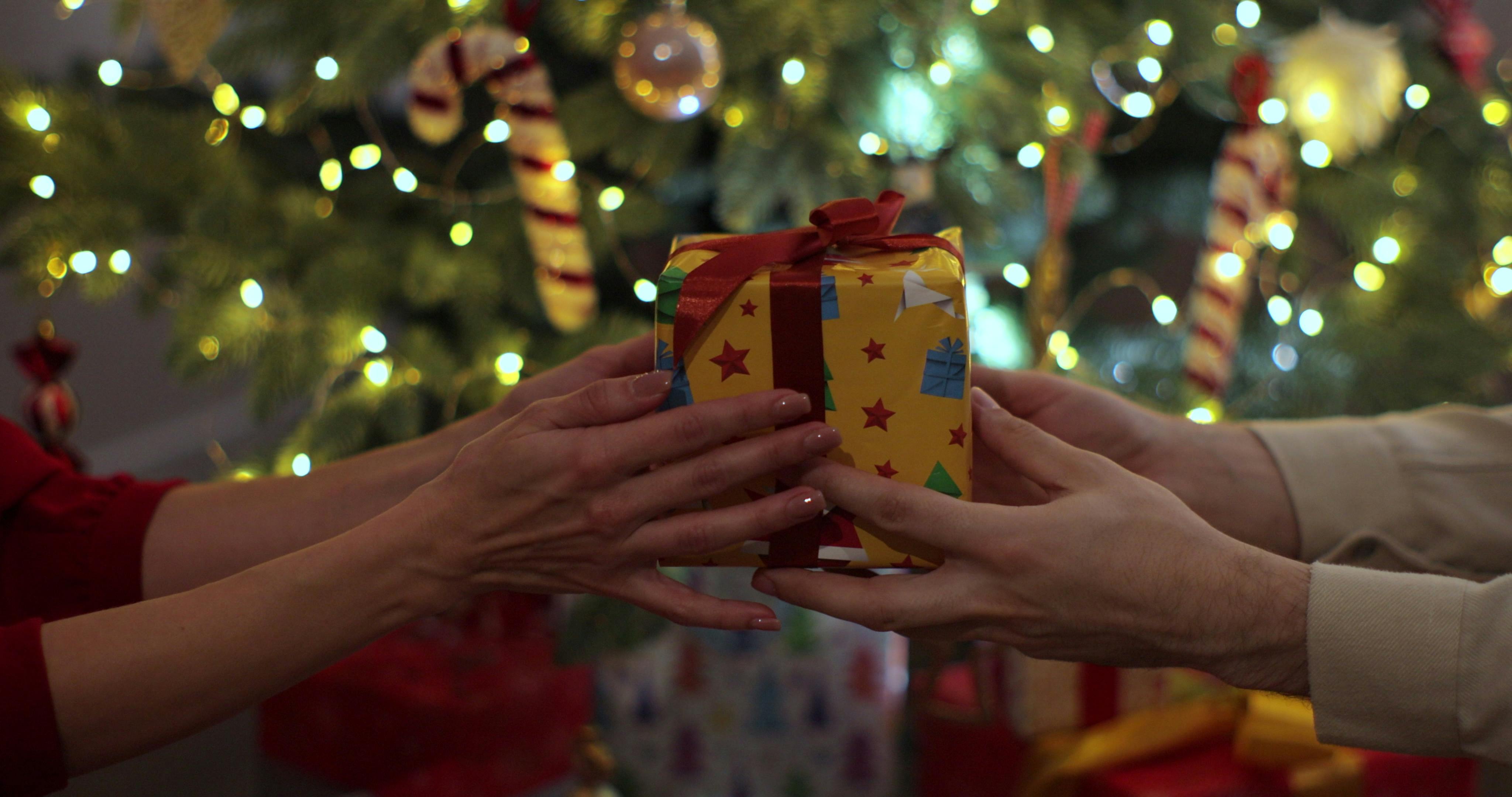 Close-up hands of man giving festive box with Christmas present to woman during holiday family party, decorating xmas tree and celebration lights