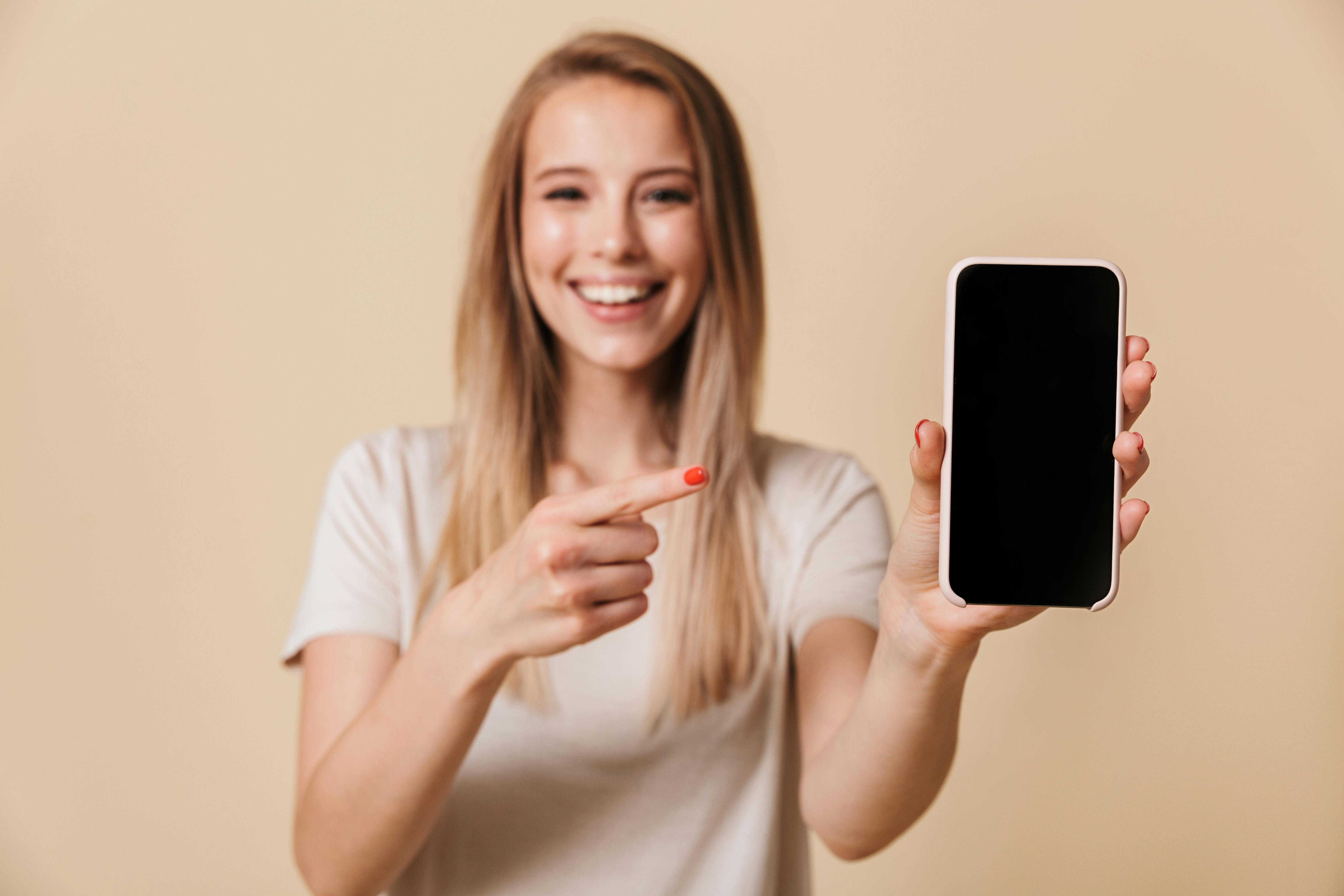 Portrait of a pretty casual girl pointing finger at blank screen mobile phone isolated over beige background