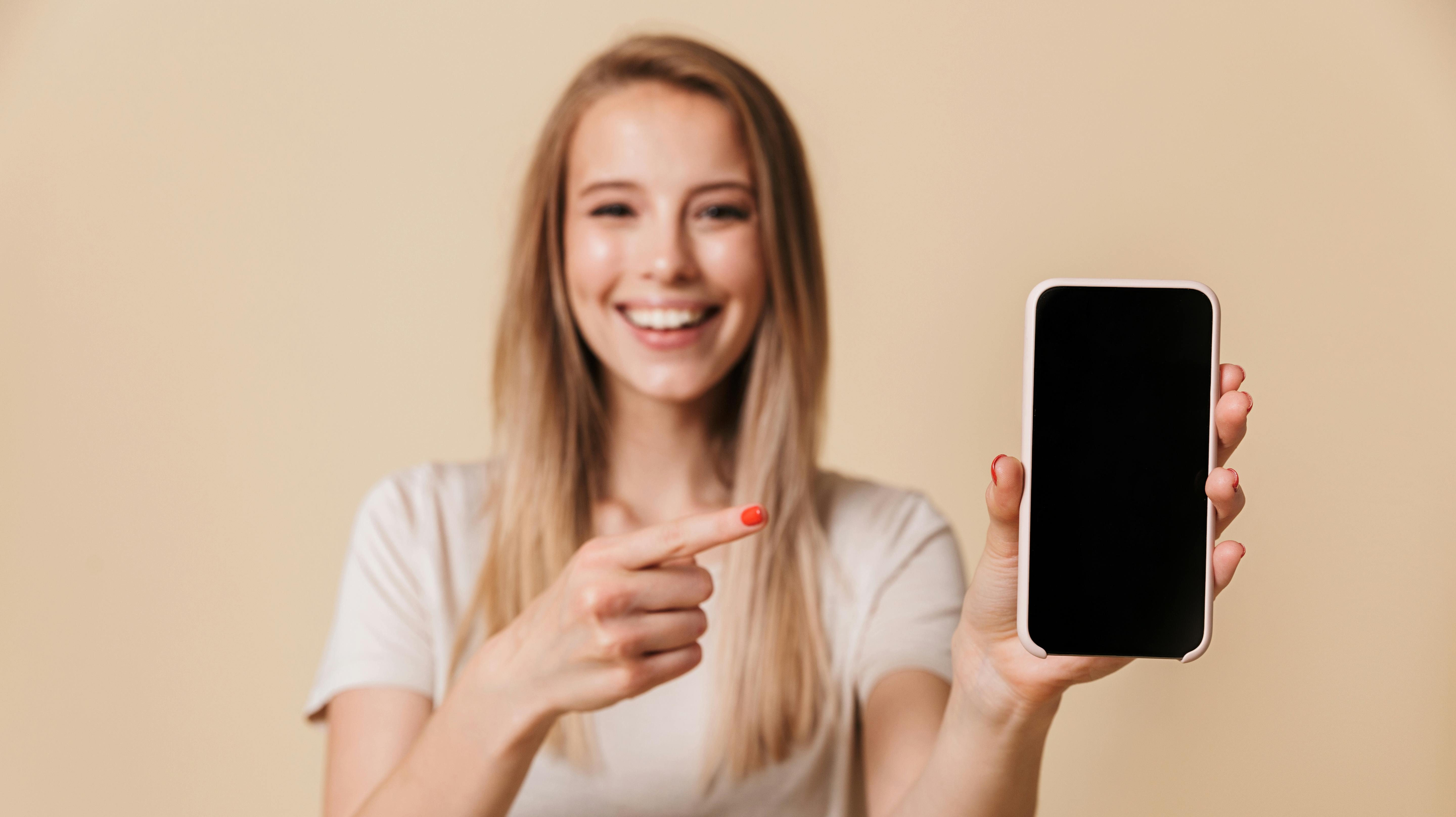 Portrait of a pretty casual girl pointing finger at blank screen mobile phone isolated over beige background