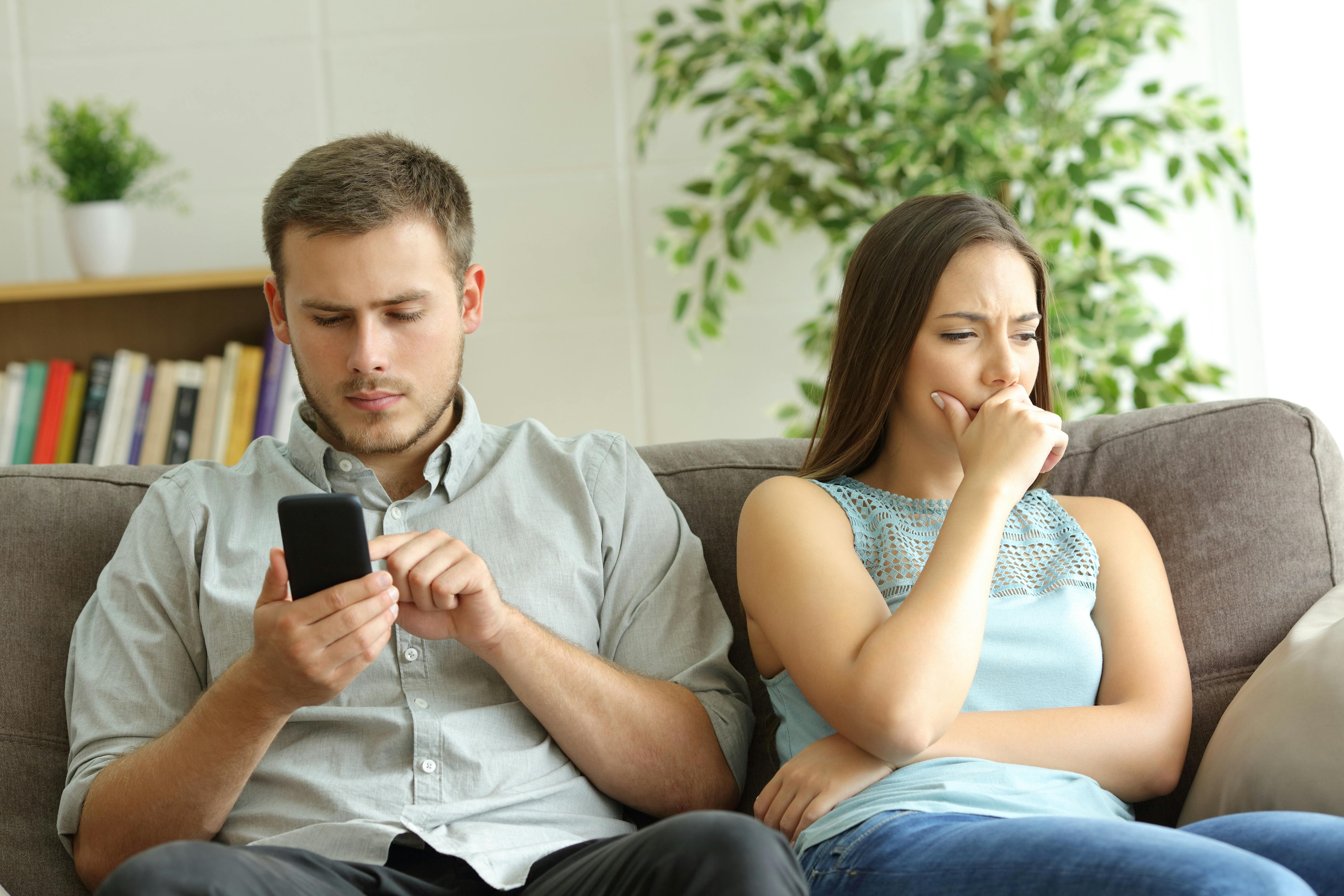 Husband addicted to smart phone watching content beside his worried wife looking down sitting on a sofa at home