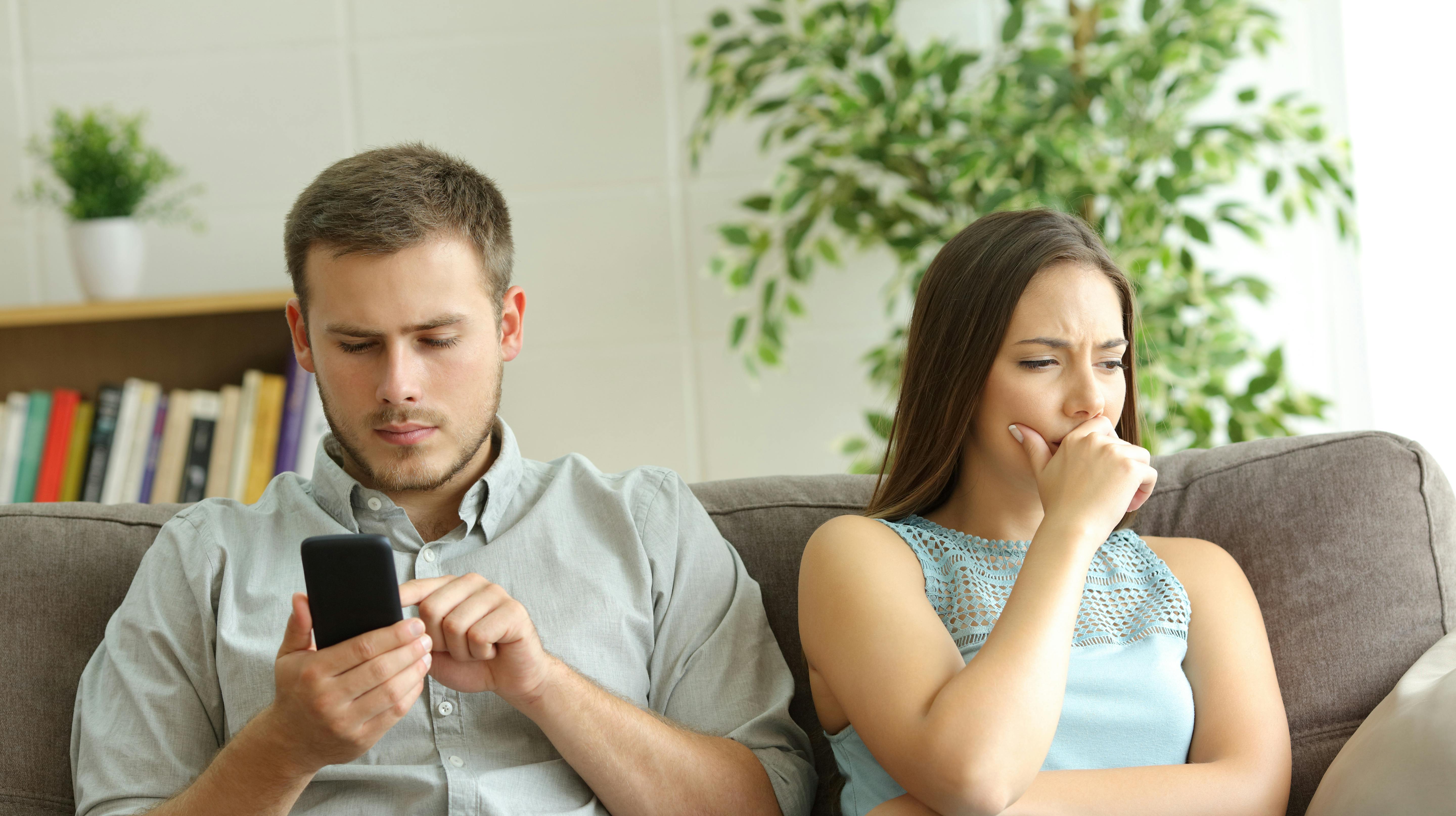 Husband addicted to smart phone watching content beside his worried wife looking down sitting on a sofa at home