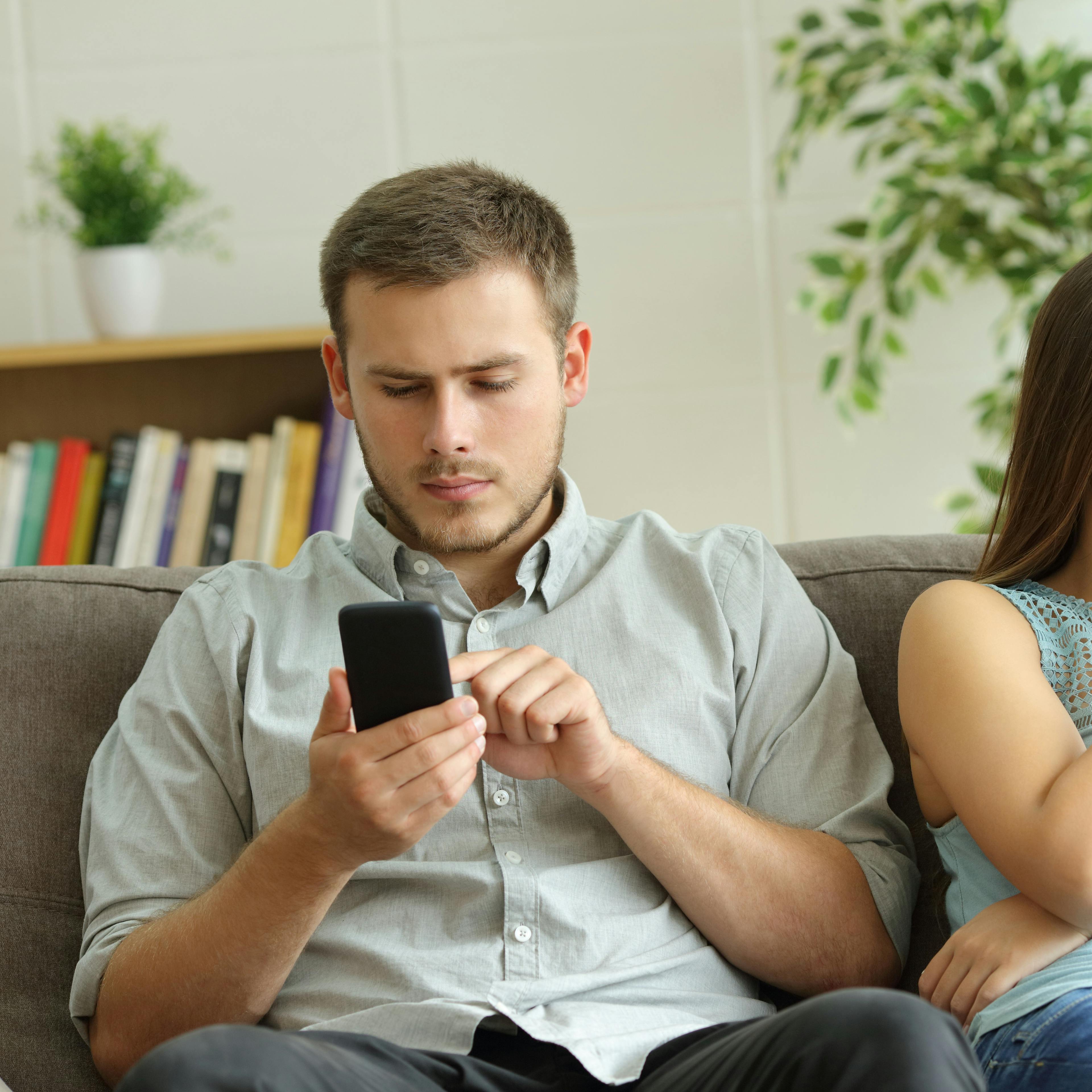 Husband addicted to smart phone watching content beside his worried wife looking down sitting on a sofa at home