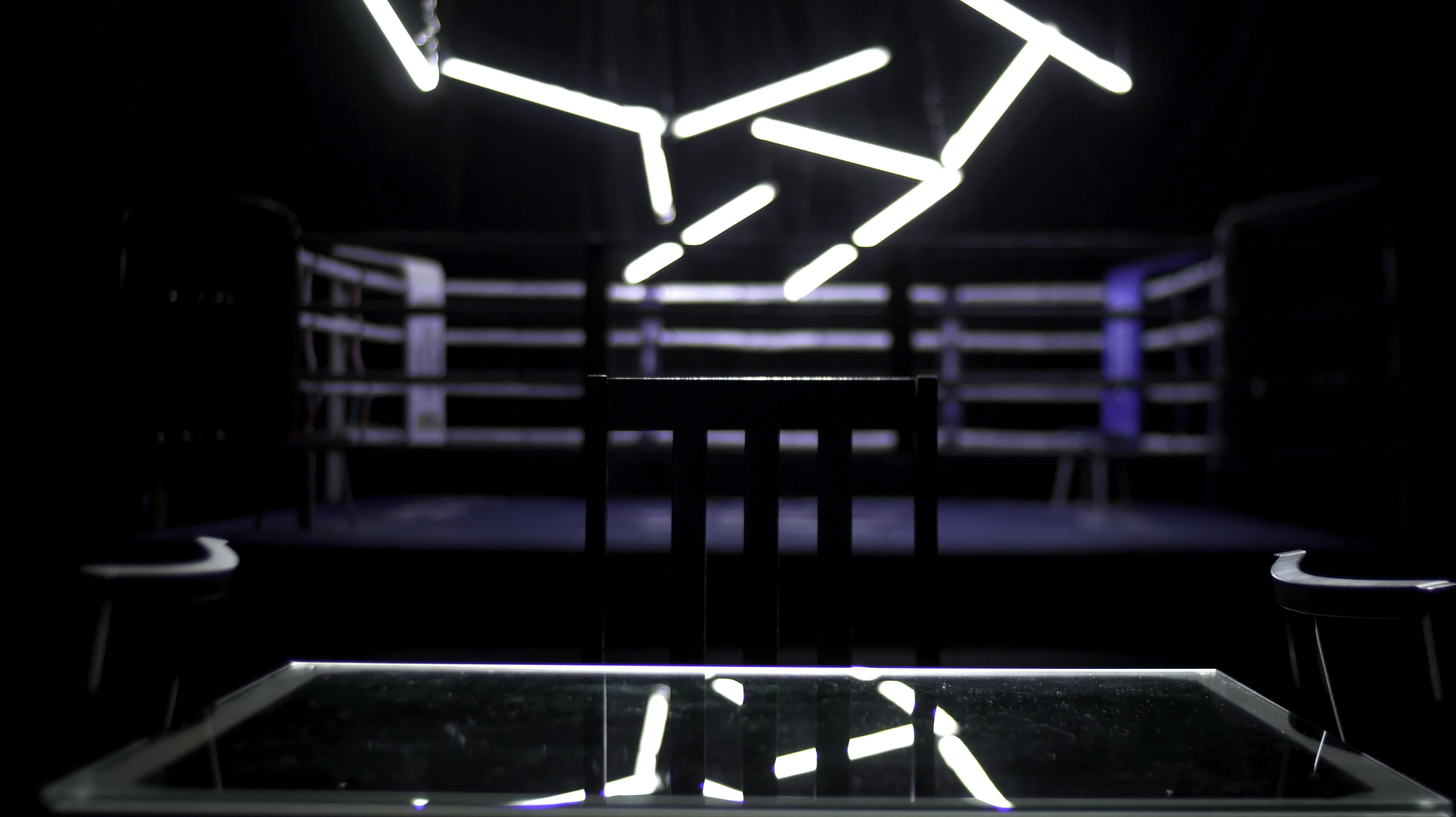 Boxing ring and two chairs with table dark background. View of a regular boxing ring surrounded by blue ropes spotlit by a spotlight. Light show around the boxing ring