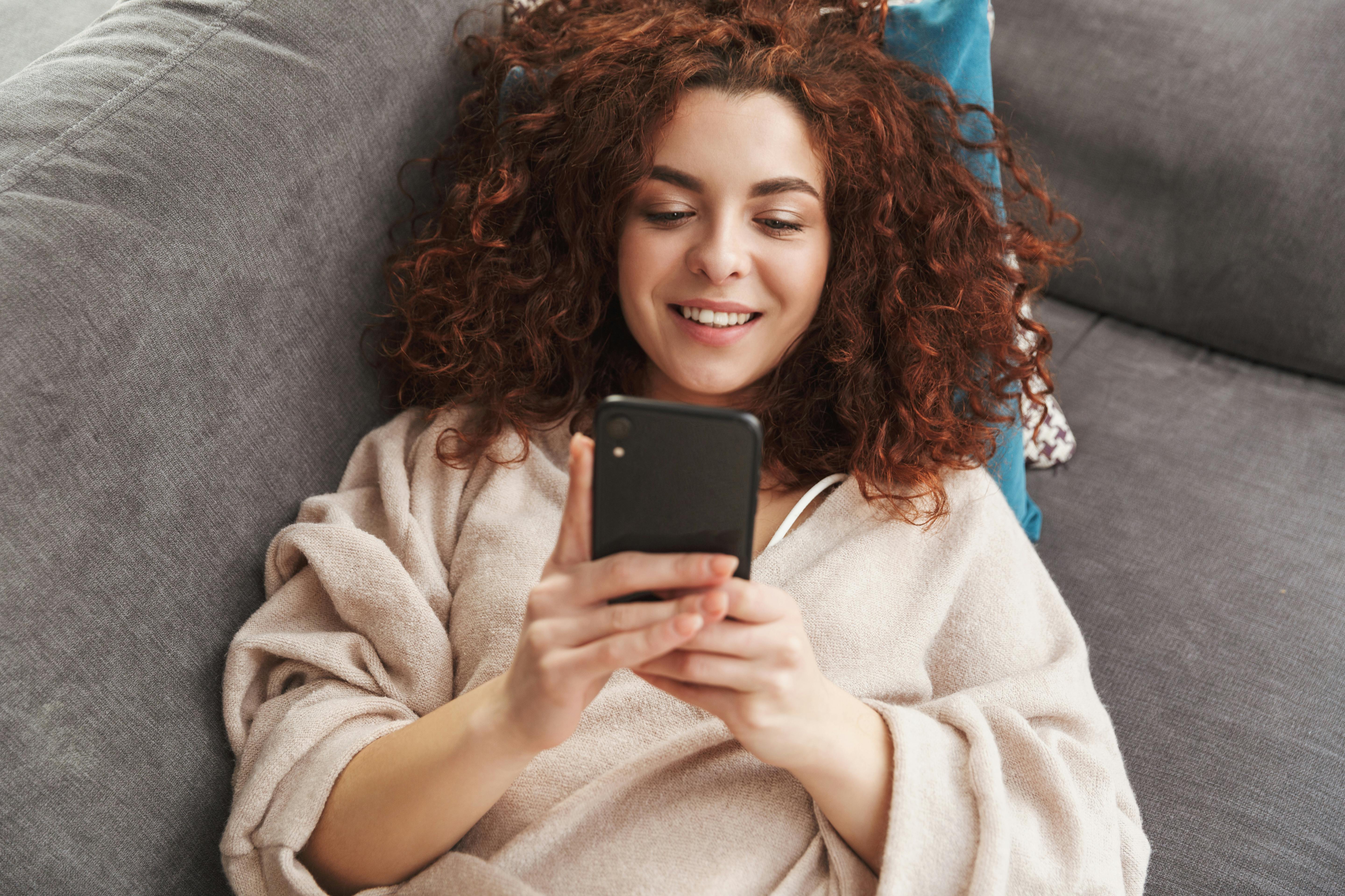 Image of curly young woman wearing house clothes lying on couch in apartment and using mobile phone