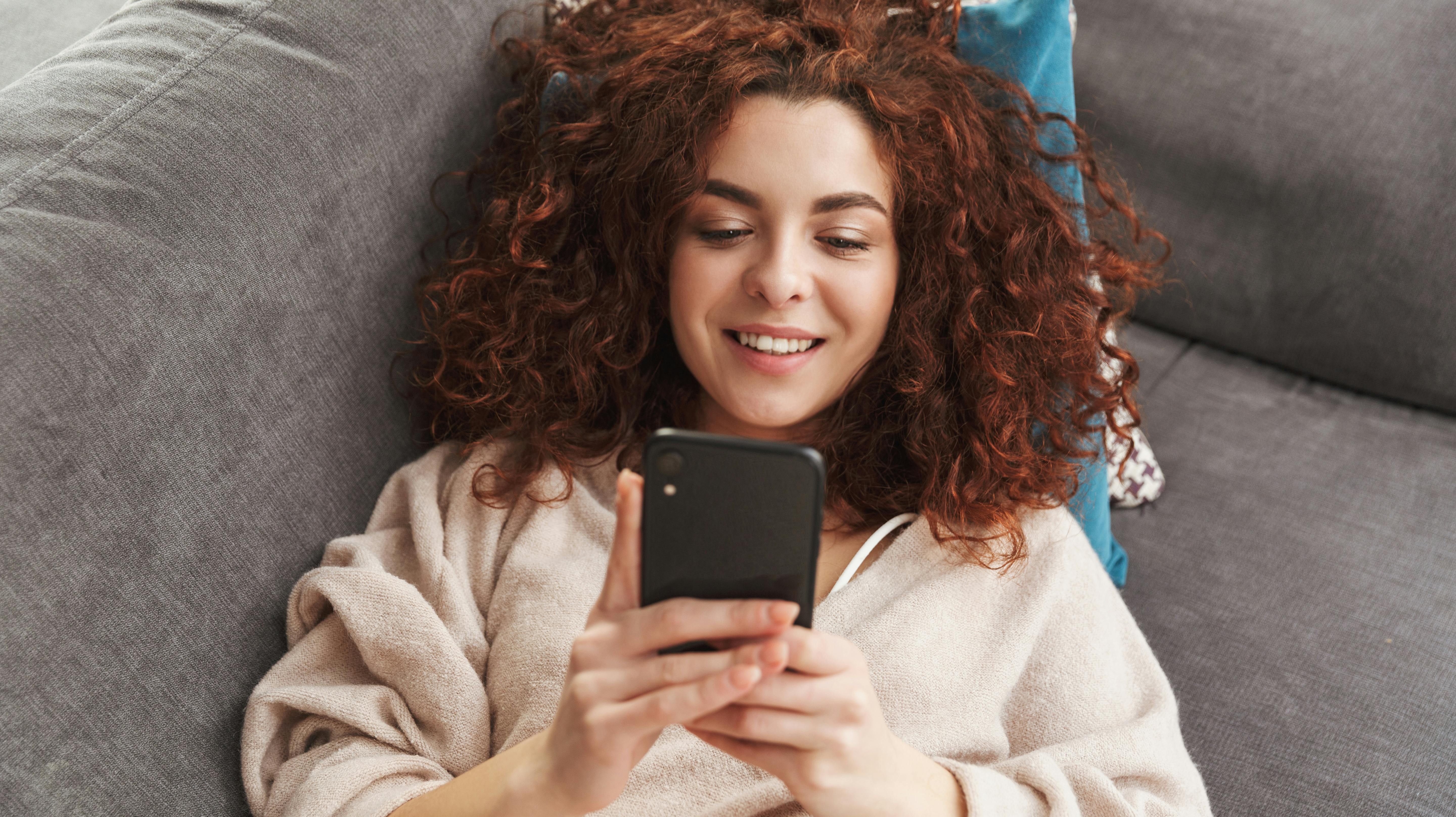 Image of curly young woman wearing house clothes lying on couch in apartment and using mobile phone