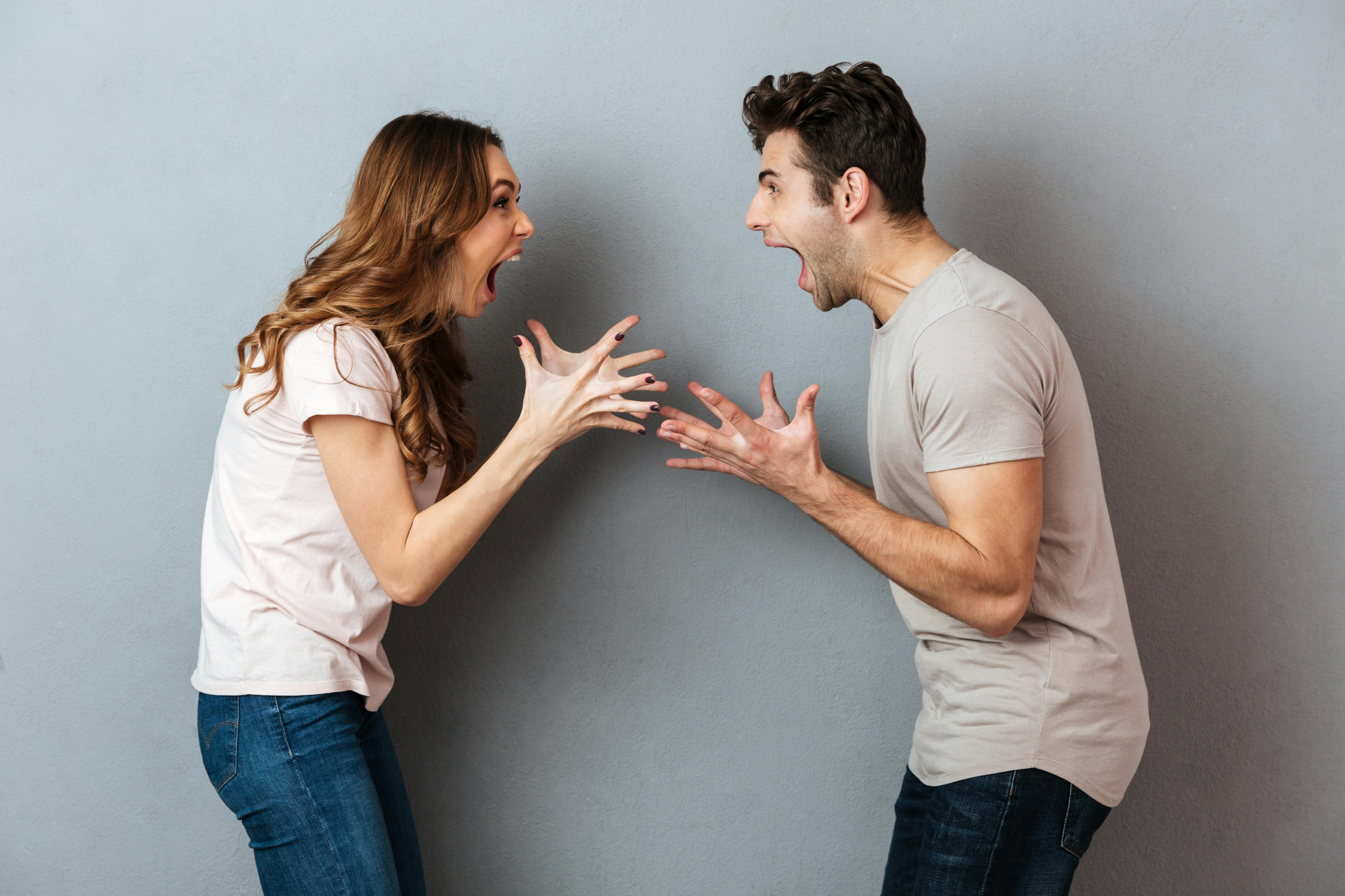 Portrait of a furious young couple having an argument while gesturing and looking at each other over gray wall