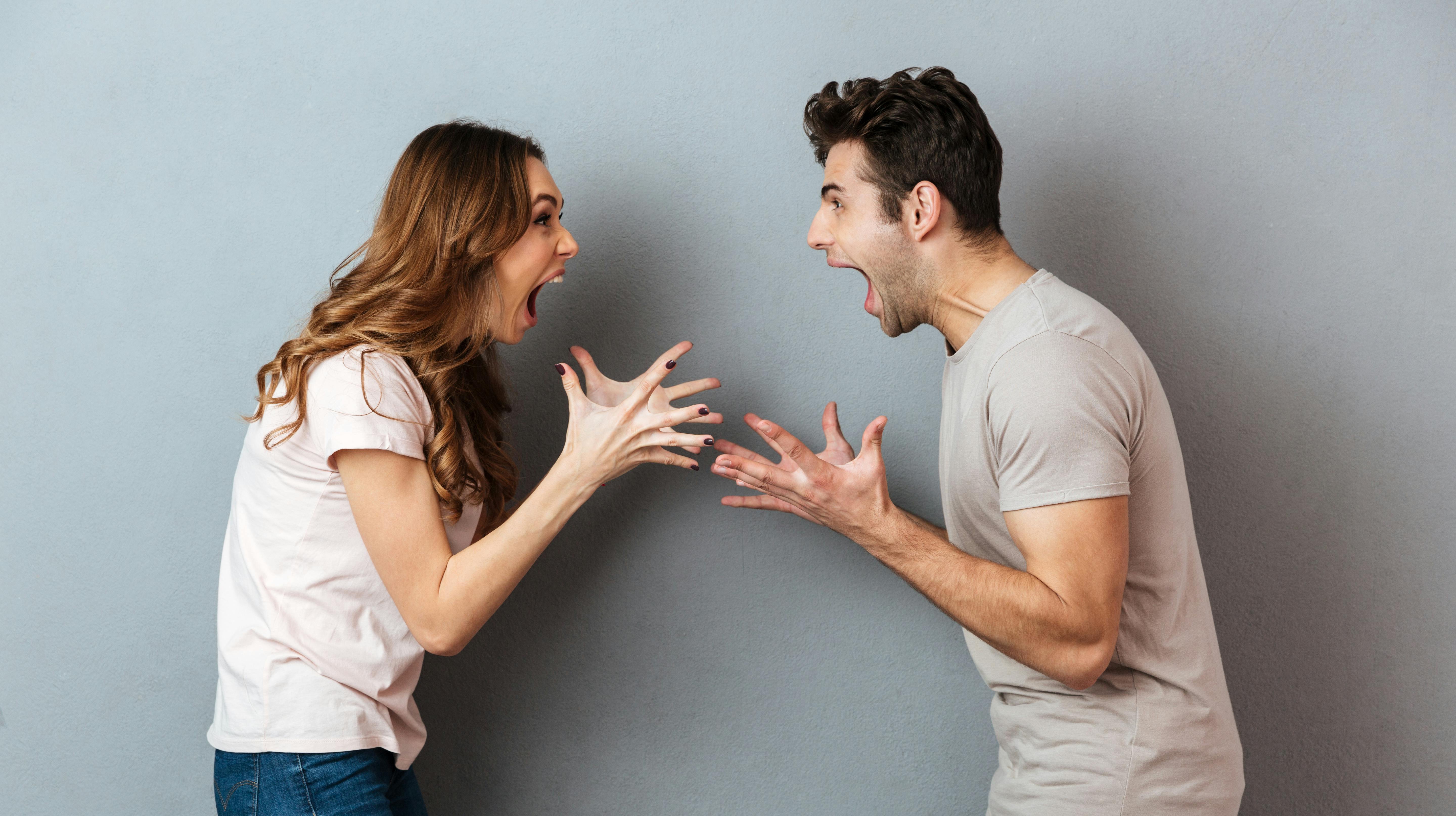 Portrait of a furious young couple having an argument while gesturing and looking at each other over gray wall