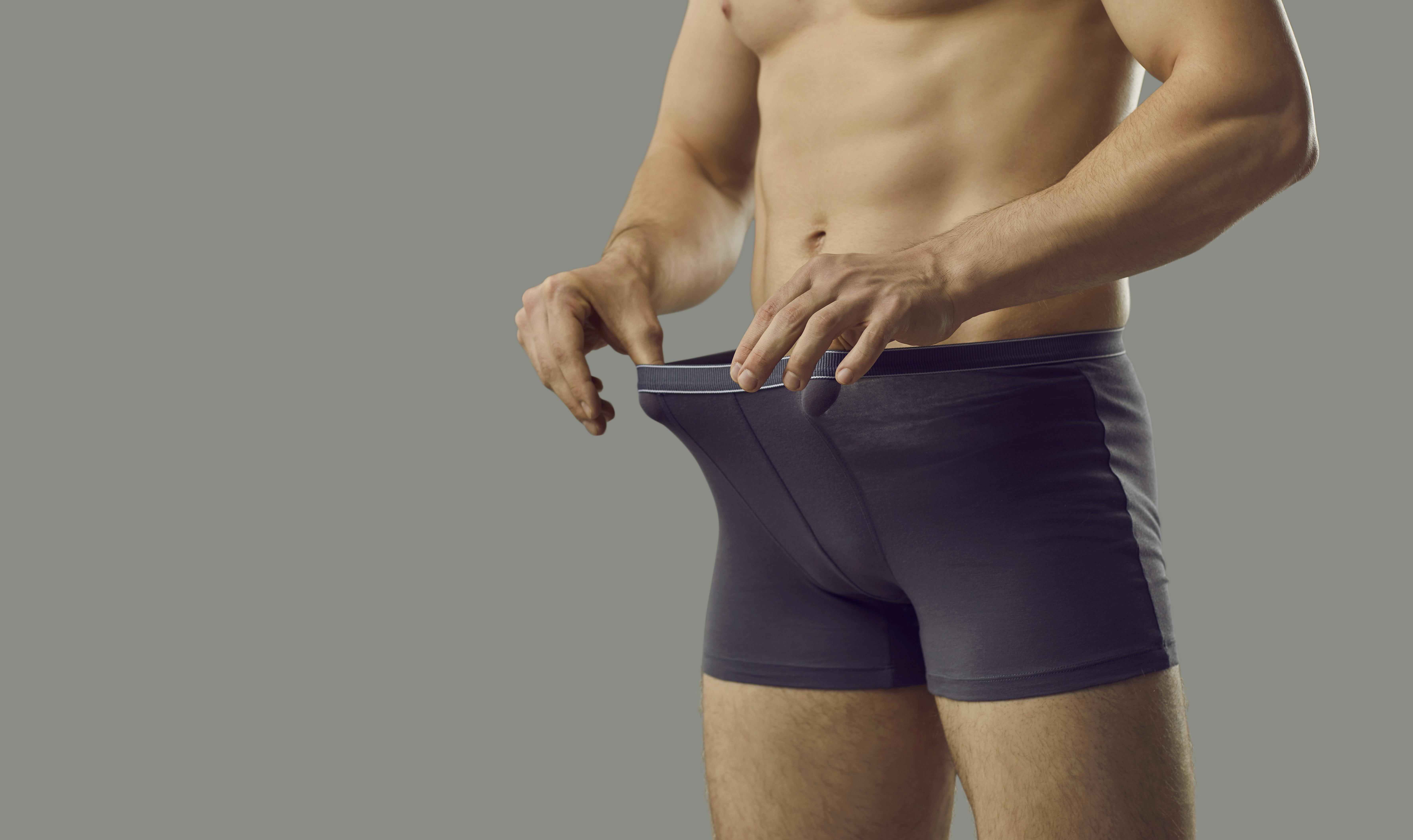 Handsome young male looks inside underpants at his private area. Shirtless attractive muscular man in underwear briefs looking at his penis standing isolated on grey background. Genital health concept