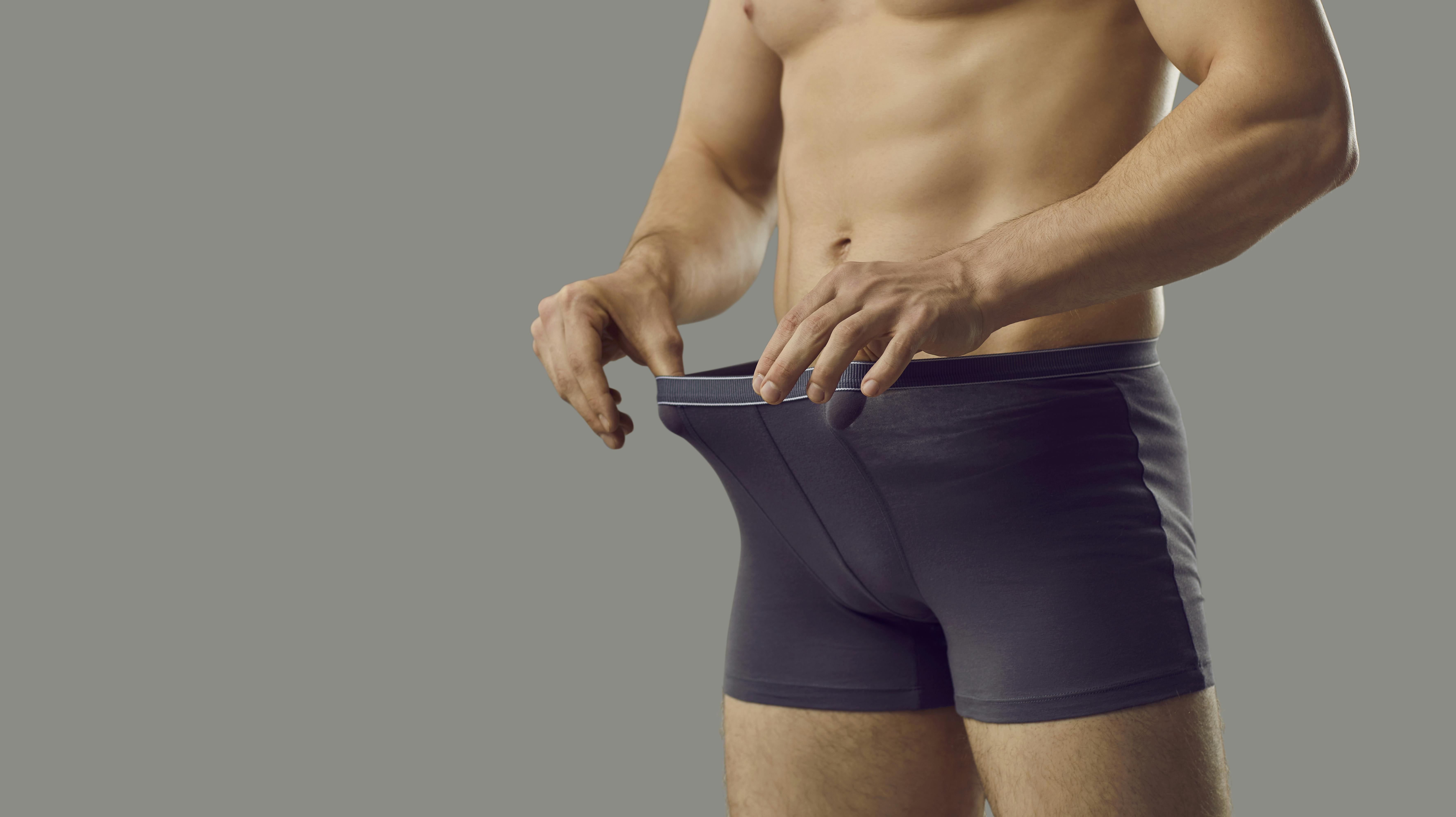 Handsome young male looks inside underpants at his private area. Shirtless attractive muscular man in underwear briefs looking at his penis standing isolated on grey background. Genital health concept