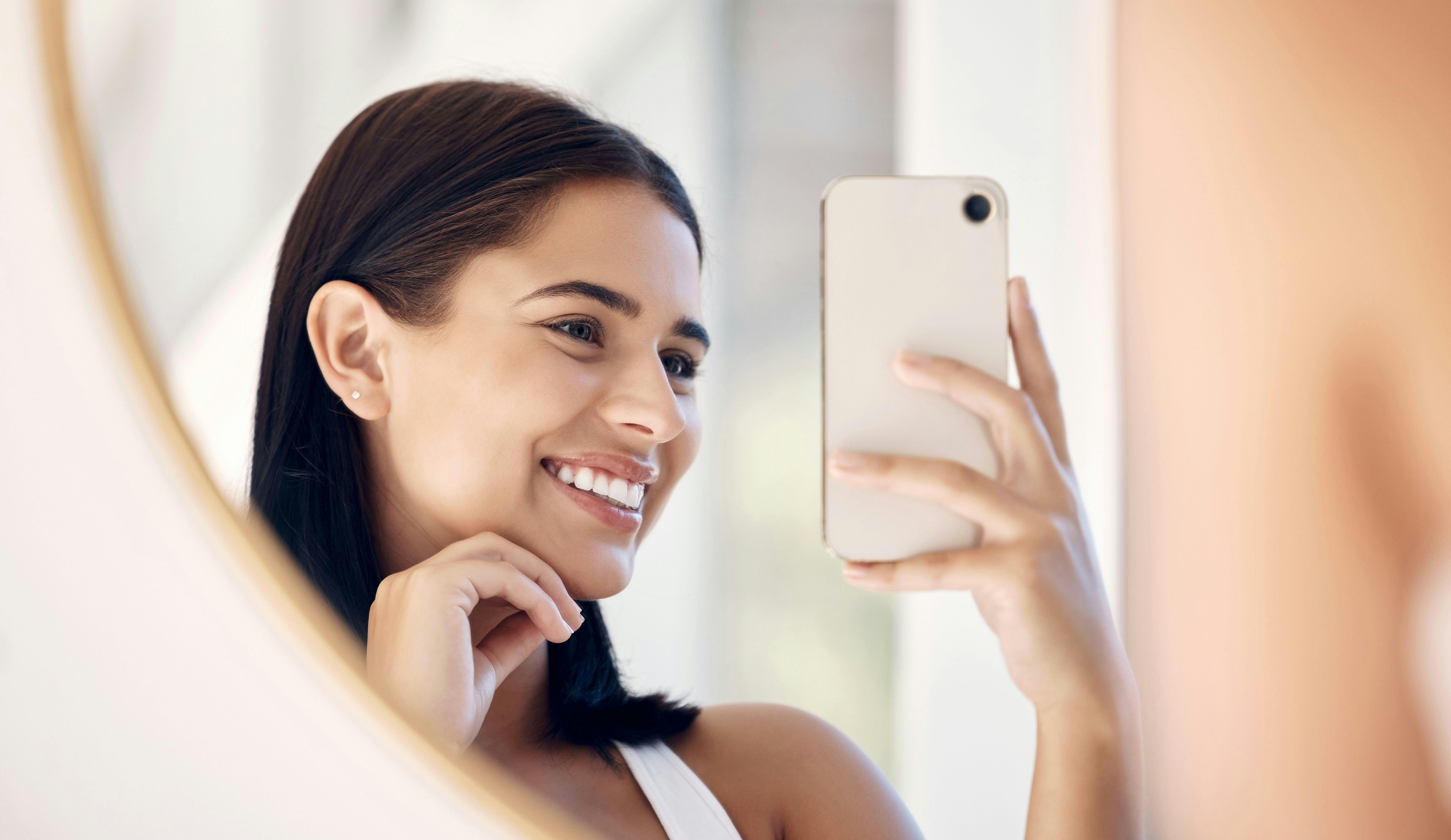 Phone, happy and selfie of woman in bathroom with smile for natural social media beauty in mirror. Reflection, happiness and skincare of wellness influencer girl with smartphone in the morning