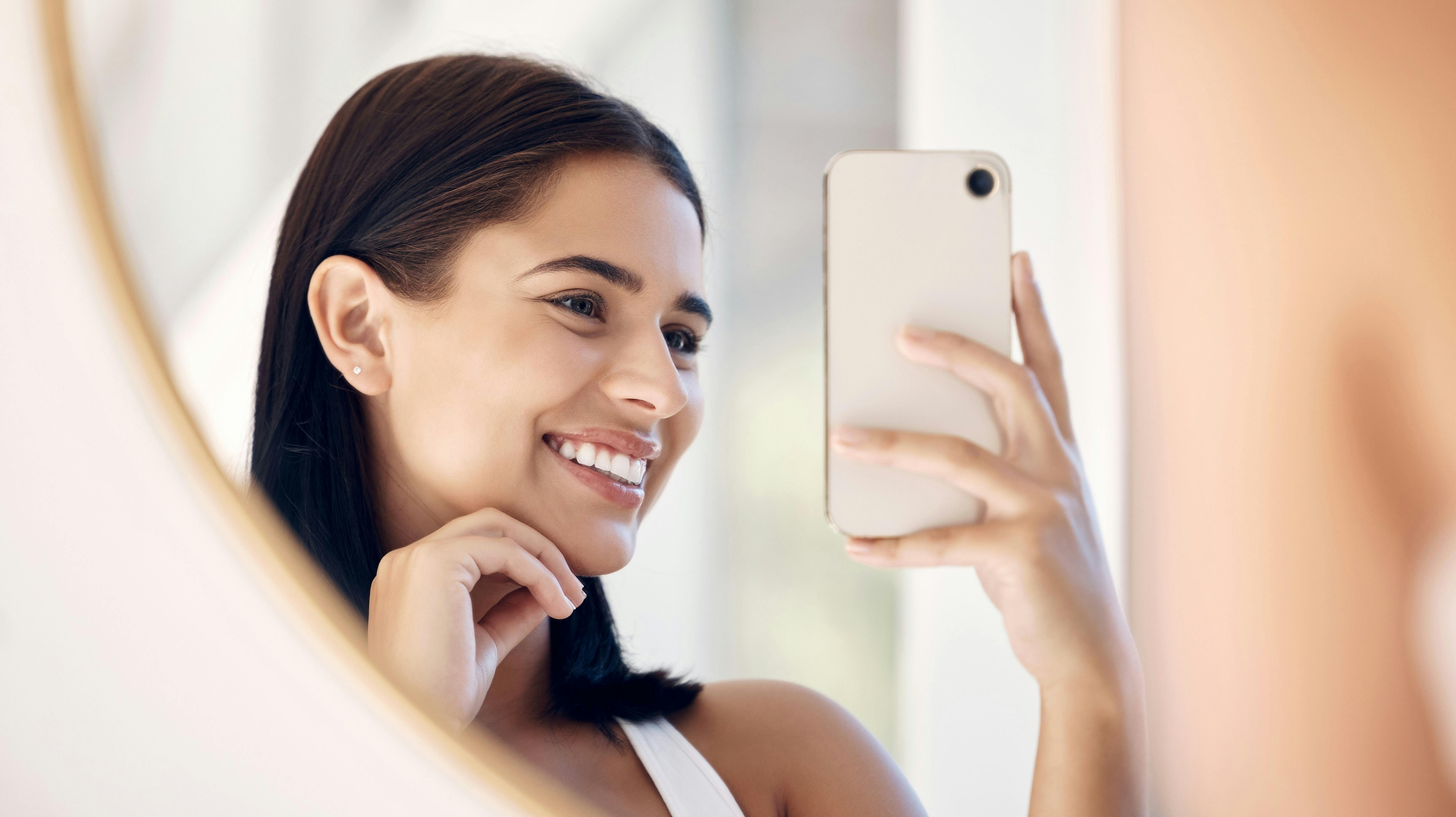 Phone, happy and selfie of woman in bathroom with smile for natural social media beauty in mirror. Reflection, happiness and skincare of wellness influencer girl with smartphone in the morning