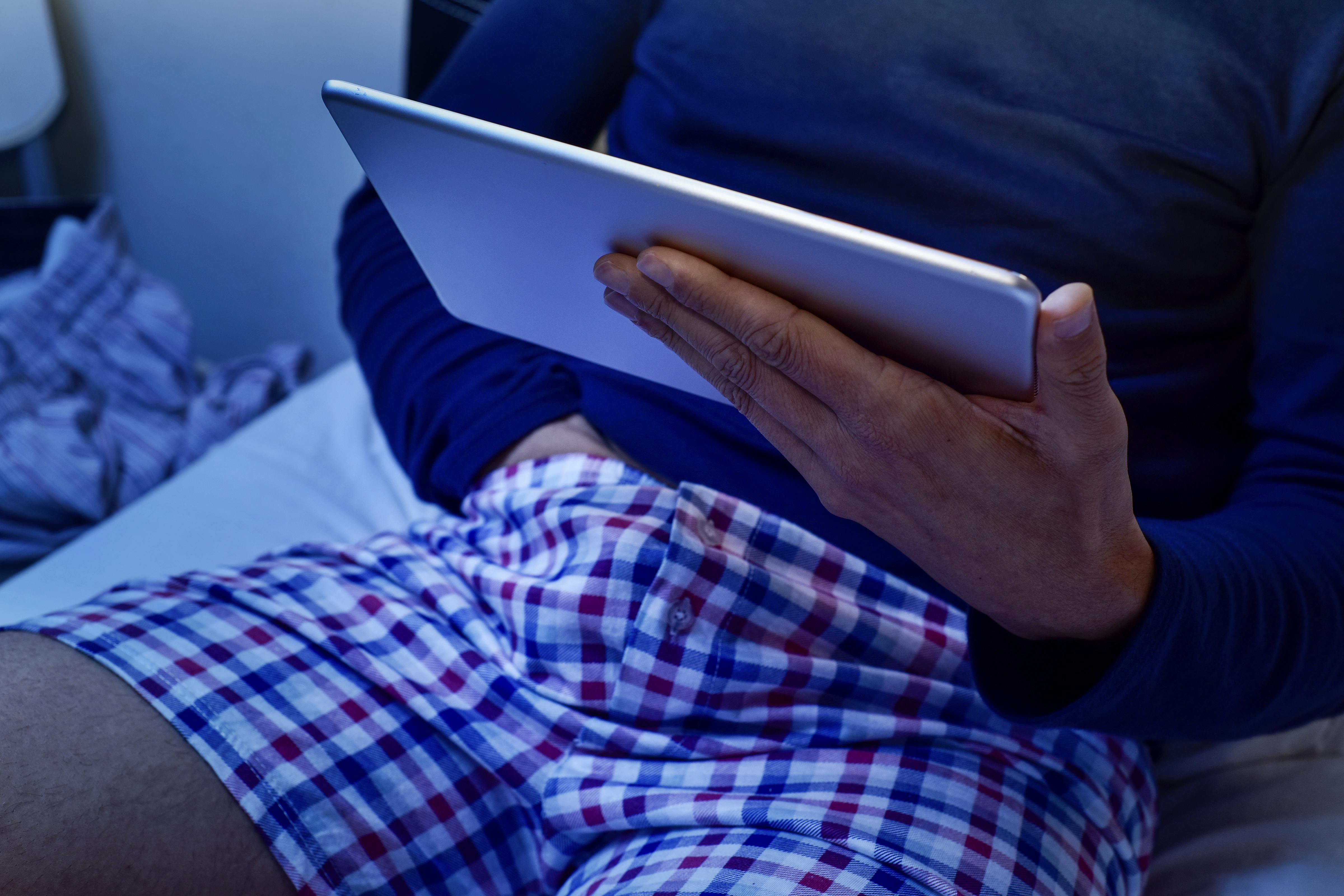 a young caucasian man lying in bed has his hand under his underwear while is watching his tablet