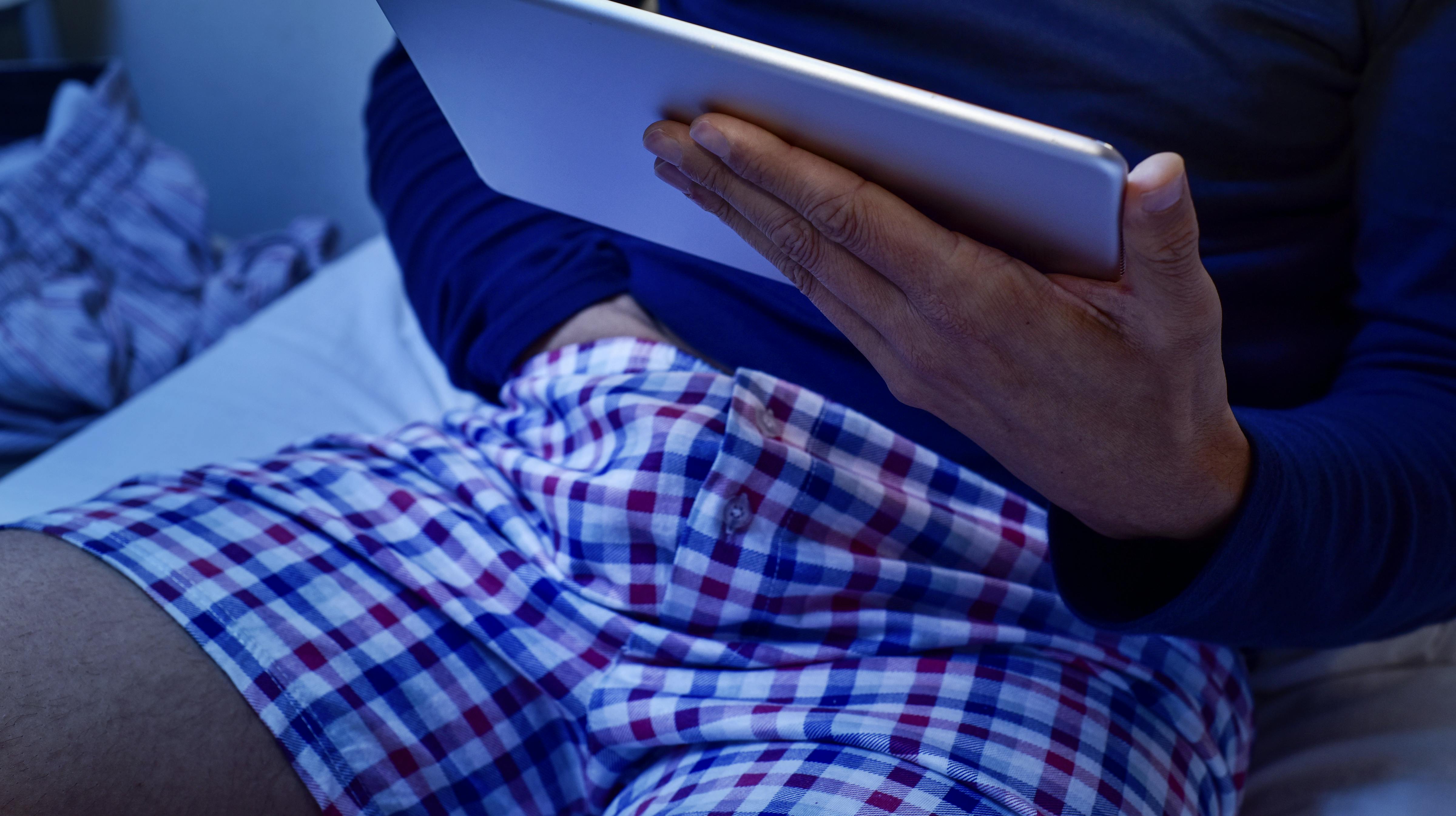 a young caucasian man lying in bed has his hand under his underwear while is watching his tablet